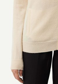 Beige stickad tröja med rund halsringning, ribbade mudd och en ficka på sidan. Slät textur, matchad med svarta byxor.