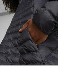 Chaqueta acolchada gris oscura con textura acolchada. Presenta una mano parcialmente en un bolsillo lateral y un acabado exterior suave y brillante.