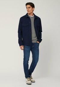 Giubbotto blu navy con bottoni sopra un maglione grigio, abbinato a jeans blu. Il giubbotto presenta due tasche frontali e una vestibilità comoda.
