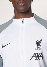 Chaqueta blanca con cremallera y detalles en gris. Presenta el logo de Nike, el emblema del Liverpool FC y la marca AXA en la parte frontal. Textura de tela suave.
