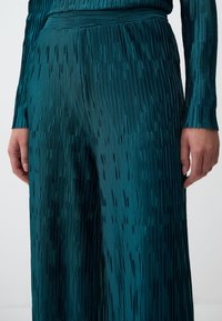 Pantaloni plissettati in teal realizzati in satin, con righe verticali, una texture liscia e una cintura larga per un fit rilassato.