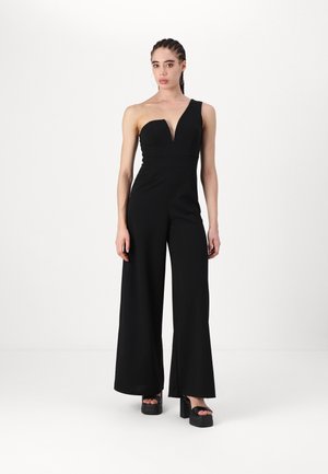 One-shoulder black jumpsuit with a plunging neckline, wide legs, and a fitted bodice. Made from smooth fabric, paired with chunky platform shoes.