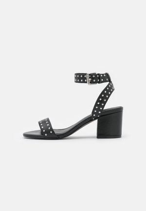 Black leather heeled sandals with an ankle strap, silver studs along the band, and a block heel for stability. Smooth texture throughout.