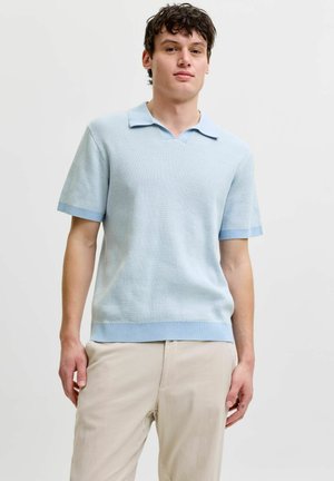 Młody mężczyzna stoi, ubrany w jasnoniebieską, krótkorękawkową dzianą koszulkę polo i beżowe spodnie, o neutralnym wyrazie twarzy na jednolitym tle.