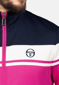 Trainingsjacke mit hohem Kragen, aus marineblauem Stoff, mit pinken und weißen horizontalen Streifen und einem Logo auf der Brust. Mit Reißverschluss.