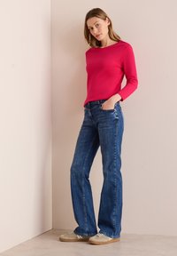 Fuchsia langarmtop gecombineerd met donkerblauwe flairjeans. Schoenen zijn lichtgrijs met witte accenten. Glad textuur, casual pasvorm.