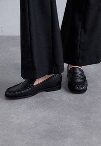 Zwarte leren loafers met een ronde neus, zachte bovenkant en decoratieve stiksels. Draag ze met losse zwarte broeken op een grijze ondergrond.