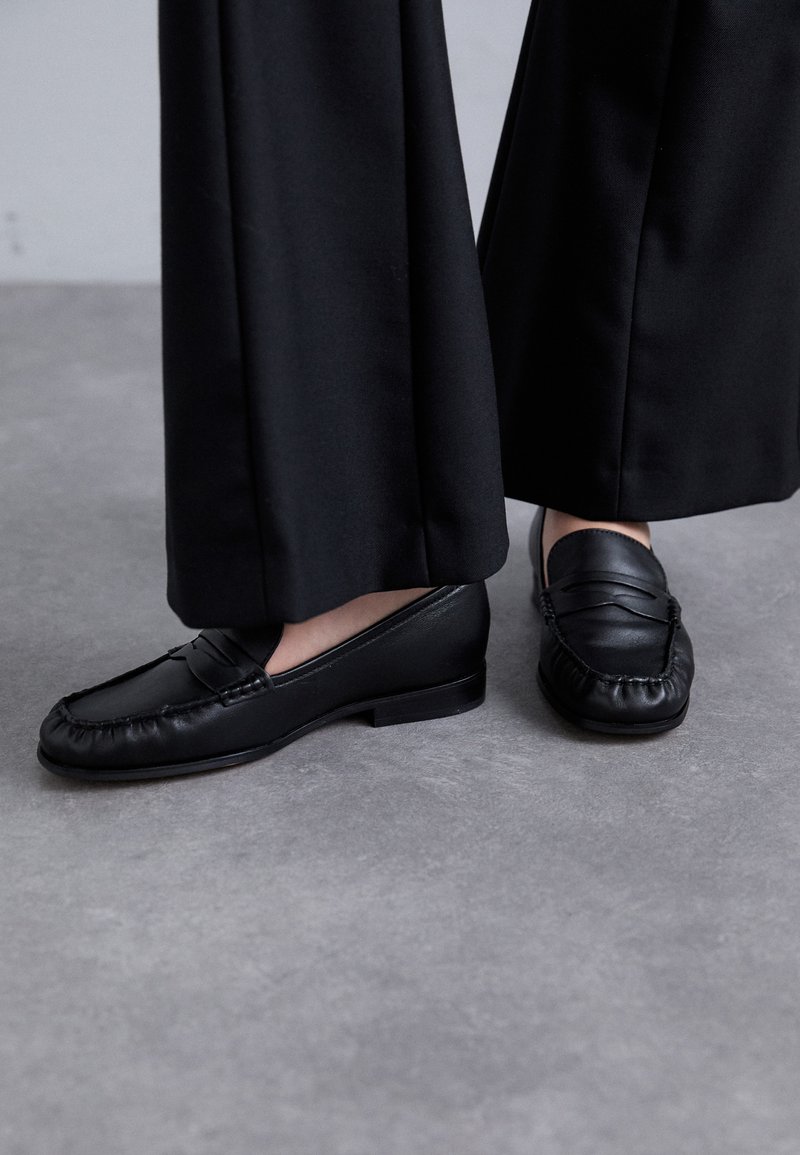 Zwarte leren loafers met een ronde neus, zachte bovenkant en decoratieve stiksels. Draag ze met losse zwarte broeken op een grijze ondergrond.