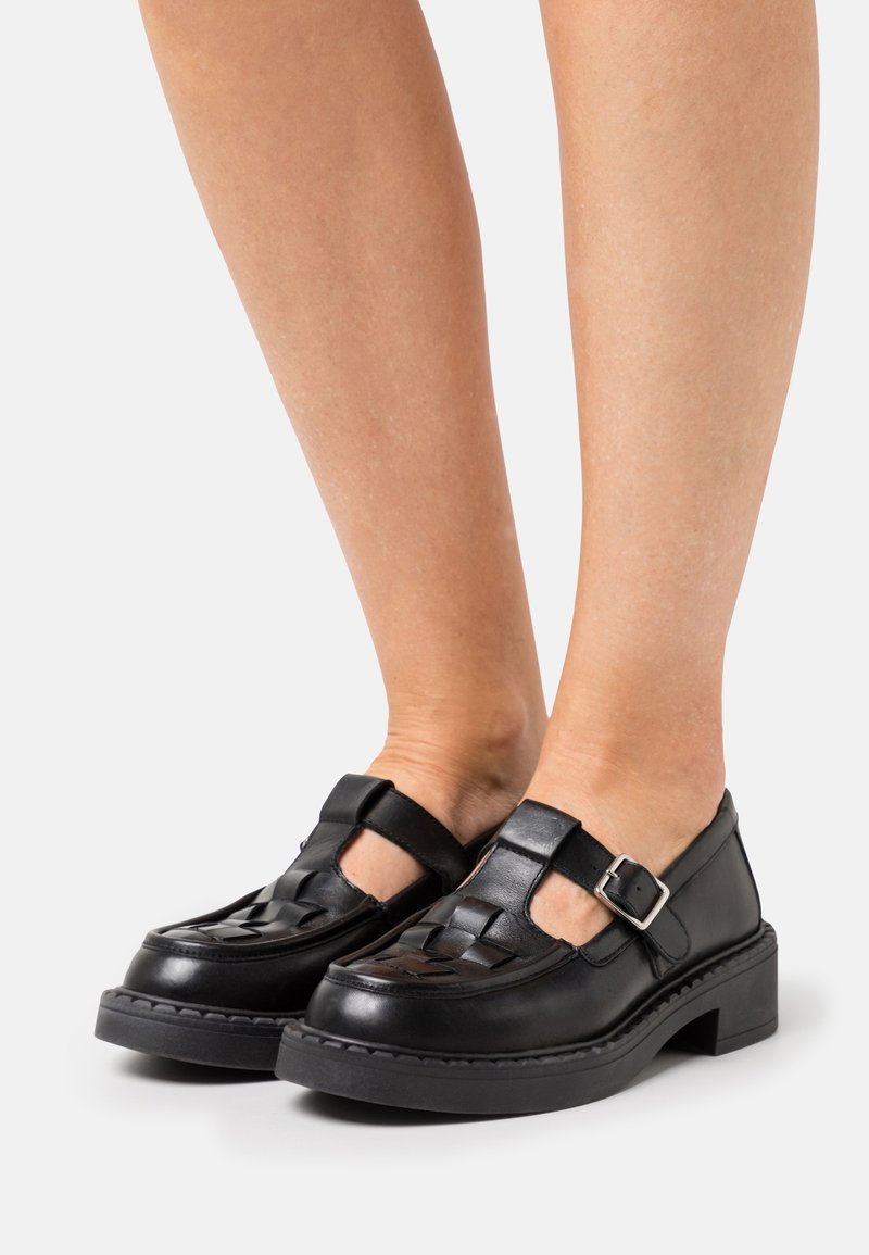 Mocassins tissés en cuir noir avec brides à boucle portés sur jambes nues sur fond blanc.