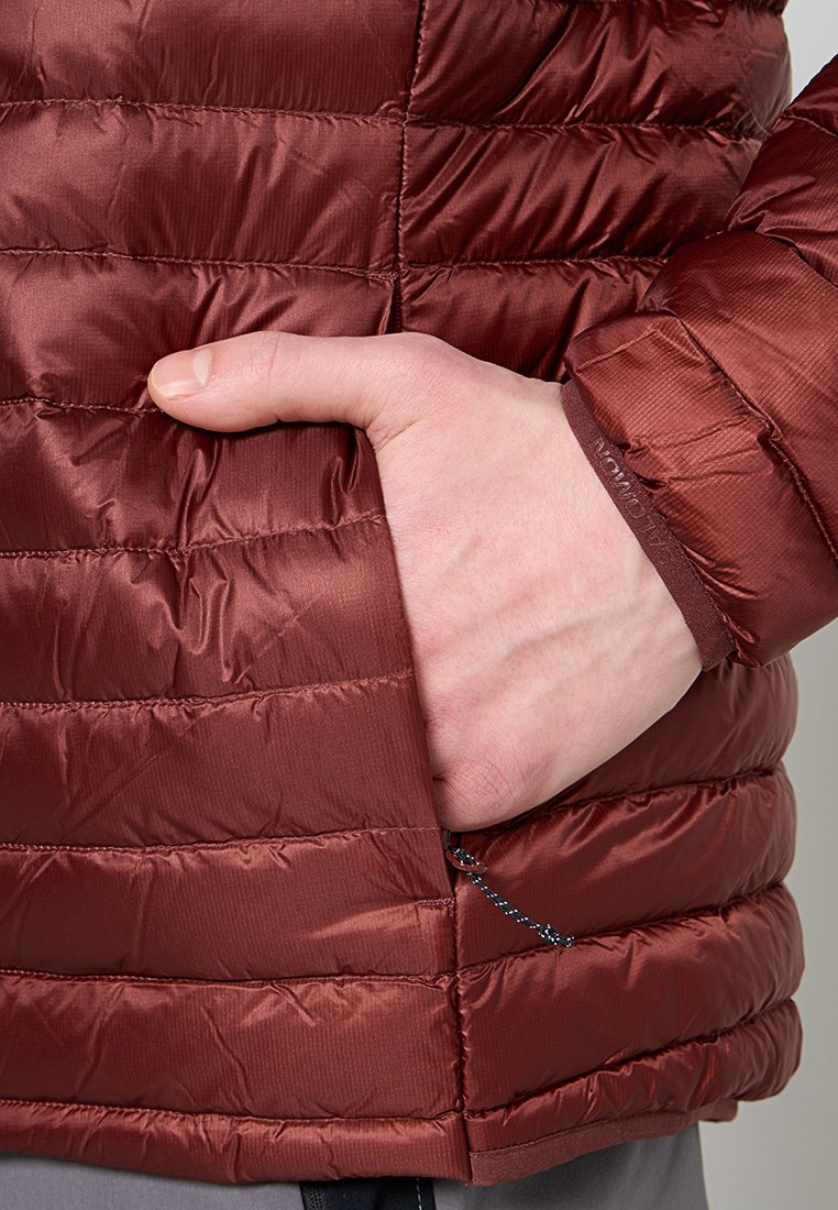 Una mano descansa dentro del bolsillo con cremallera de una chaqueta acolchada brillante de color óxido, mostrando la costura detallada de la tela y un tirador de cremallera con cordón.
