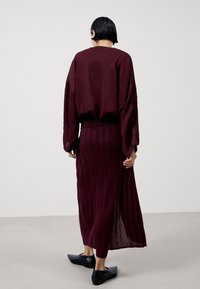 Viininpunainen neulepusero lankeavilla harteilla, yhdistetty samansävyiseen, lattiaan ulottuvaan laskostettuun hameeseen. Asussa on teksturoitu pinta ja sivuhalkio.