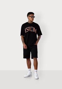 T-shirt noir avec "ESTEEM" imprimé en corail, associé à un short noir. Porté avec des baskets blanches et des lunettes de soleil, représentant une tenue décontractée.