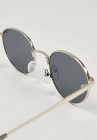 Okrągłe okulary przeciwsłoneczne z ciemnymi soczewkami, cienkimi srebrnymi metalowymi oprawkami, regulowanymi noskami oraz prostymi zausznikami o wypolerowanym wykończeniu.