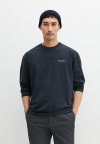 Marineblauer langärmliger Pullover mit Rundhalsausschnitt; weicher Stoff; mit Logo-Stickerei auf der linken Brust; kombiniert mit grauer Hose und einer dunklen Mütze.