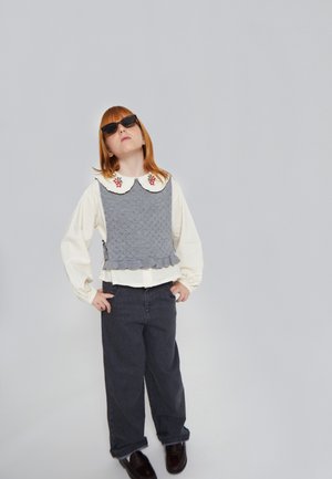 Chaleco gris de punto con dobladillo con ondas sobre una blusa blanca, acentuado por un gran cuello y adornos florales, combinado con pantalones oscuros y gafas de sol.