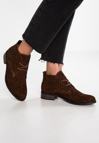 Stivali alla caviglia in suede marrone con una fantasia maculata, dotati di chiusura con lacci e tacco basso impilato, abbinati a jeans neri sfilacciati.