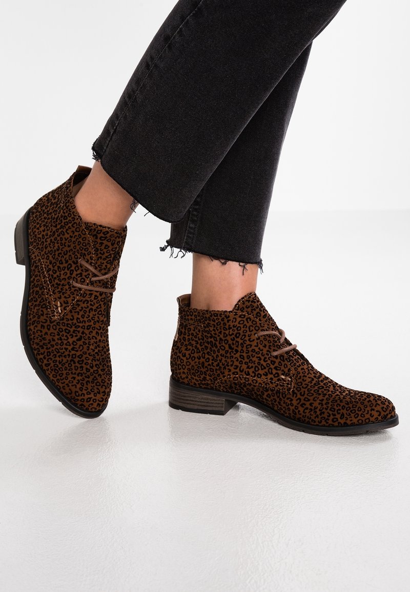 Stivali alla caviglia in suede marrone con una fantasia maculata, dotati di chiusura con lacci e tacco basso impilato, abbinati a jeans neri sfilacciati.