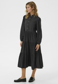 Woman with wavy hair wearing a long-sleeve, button-front, tiered black midi dress and black loafers, standing and looking to the side.