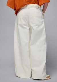 Wide-leg white cotton trousers with back pockets, a belt loop, and a small logo on the right side. Worn with an orange textured top.