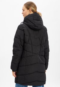 Vrouw met blond haar in een lage paardenstaart, gekleed in een zwarte gewatteerde winterjas met capuchon en blauwe spijkerbroek, kijkt naar links.