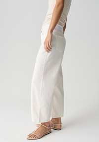 Persoon in een crèmekleurige tanktop, hoge taille, wijde witte broek en beige platte sandalen met bandjes, zijaanzicht op een lichte achtergrond.