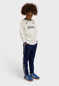 Child with curly hair looks down wearing a white long-sleeve sports jersey, navy track pants with white stripes, and blue sneakers.