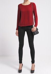 Rote Bluse mit langen Ärmeln, kombiniert mit schwarzen Skinny-Jeans und schwarzen High-Heels. Die Accessoires umfassen eine kleine schwarze Handtasche mit einer Kettenriemen.