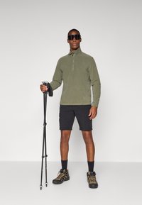 Olivgrüner Fleece-Pullover mit Reißverschlusskragen, schwarze Shorts, schwarze Wanderschuhe mit orangefarbenen Akzenten und Trekkingstöcke. Glatte Textur.