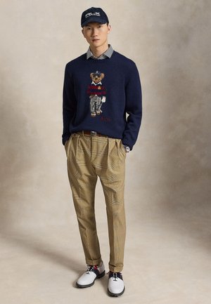 Homme portant un pull bleu marine avec un motif ours, une chemise rayée, un pantalon plissé beige, des chaussures blanches et une casquette bleu marine, debout les mains dans les poches.