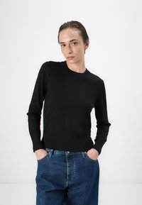 Svart langermet genser med rund hals og ribbestrikkede mansjetter, kombinert med blå høyt liv-jeans, har en jevn tekstur.