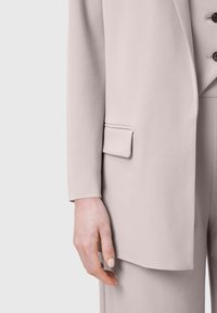 Gaiši rozā žakete no gludas auduma; ar vienu atloku kabatu un trim pogu rotājumiem priekšpusē, ar pieguļošām piedurknēm.