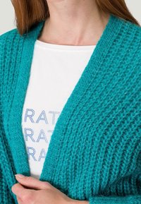 Türkiser Strickcardigan mit einem strukturierten, lockeren Gewebedesign. Offene Vorderseite und weite Ärmel, über einem weißen T-Shirt mit bedruckten Details getragen.