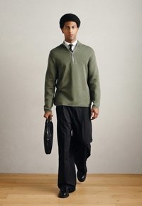 Πράσινο πουλόβερ με φερμουάρ, μαύρο επίσημο παντελόνι και παπούτσια. Κρατά μια μαύρη τσάντα χειρός. Στέκεται σε ξύλινο πάτωμα με ουδέτερο φόντο.