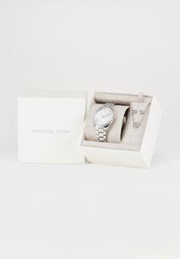 Zilveren horloge met metalen band, bijpassende ketting en oorbellen set in een witte Michael Kors doos met zachte beige binnenkant.