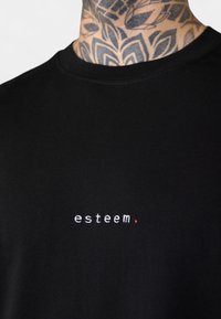 Schwarzes T-Shirt aus Baumwolle mit weiß gesticktem Text "esteem." und einem roten Punktakzent. Kurze Ärmel und Rundhalsausschnitt.