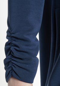 Navyblaues Oberteil mit Ärmeln, das mit gerafftem Rüschendetail versehen ist, aus weichem, elastischem Stoff gefertigt. Glatte Textur mit einer figurbetonten Silhouette.