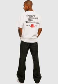 Hvid t-shirt med sort og rød grafisk tekst på ryggen, parret med mørke jeans. Det samlede look har en afslappet pasform og en uformel stil.