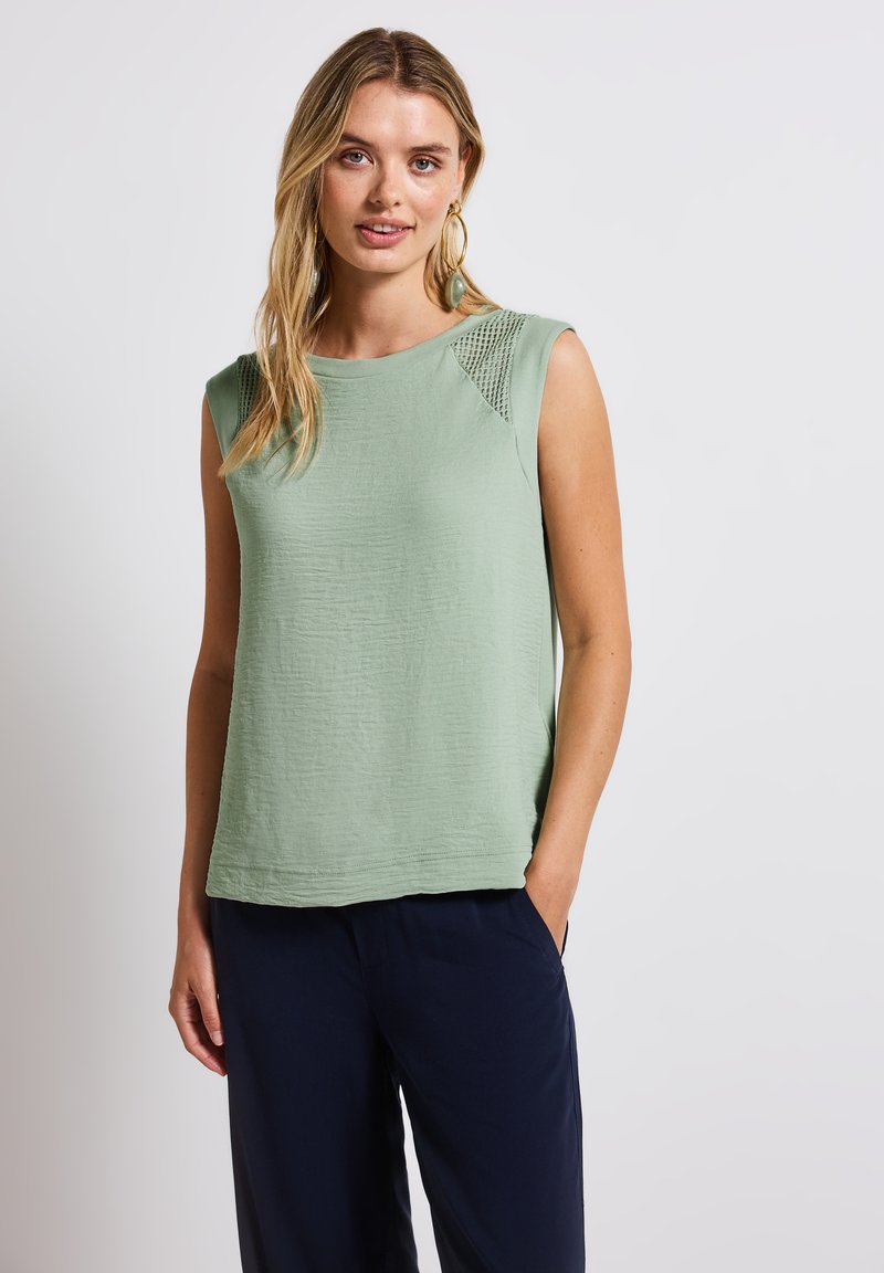 Ljust grön ärmlös topp med en texturerad yta och meshdetalj på axlarna, kombinerad med mörkblå lösa byxor.