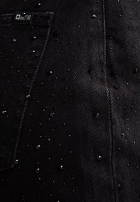 Tela de mezclilla negra con adornos de cuentas. Presenta un bolsillo y una etiqueta visible. La superficie tiene un aspecto texturizado con cuentas de diferentes tamaños.