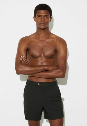 Czarne kąpielówki o gładkiej fakturze, z elastycznym paskiem w talii i złotym guzikiem jako detalem. Model o atletycznej sylwetce stoi z założonymi ramionami.