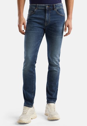 Blauwe spijkerbroek met een slim fit, vijf zakken en lichte vervaging. G gedragen met witte mesh sneakers.