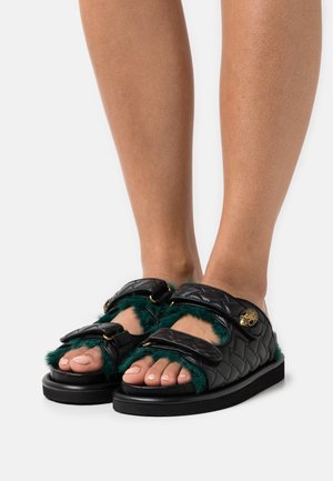Pieds portant des sandales noires matelassées avec une doublure en fourrure verte et des détails à boucle dorée, présentés sur un fond blanc.