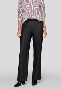 Frau trägt schwarze weite Hosen, schwarze Stiefel mit Absatz und ein lila-weiß gestreiftes Hemd mit hochgekrempelten Ärmeln.