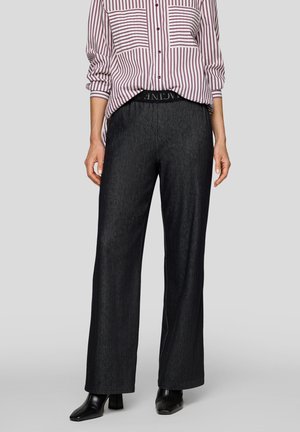 Frau trägt schwarze weite Hosen, schwarze Stiefel mit Absatz und ein lila-weiß gestreiftes Hemd mit hochgekrempelten Ärmeln.