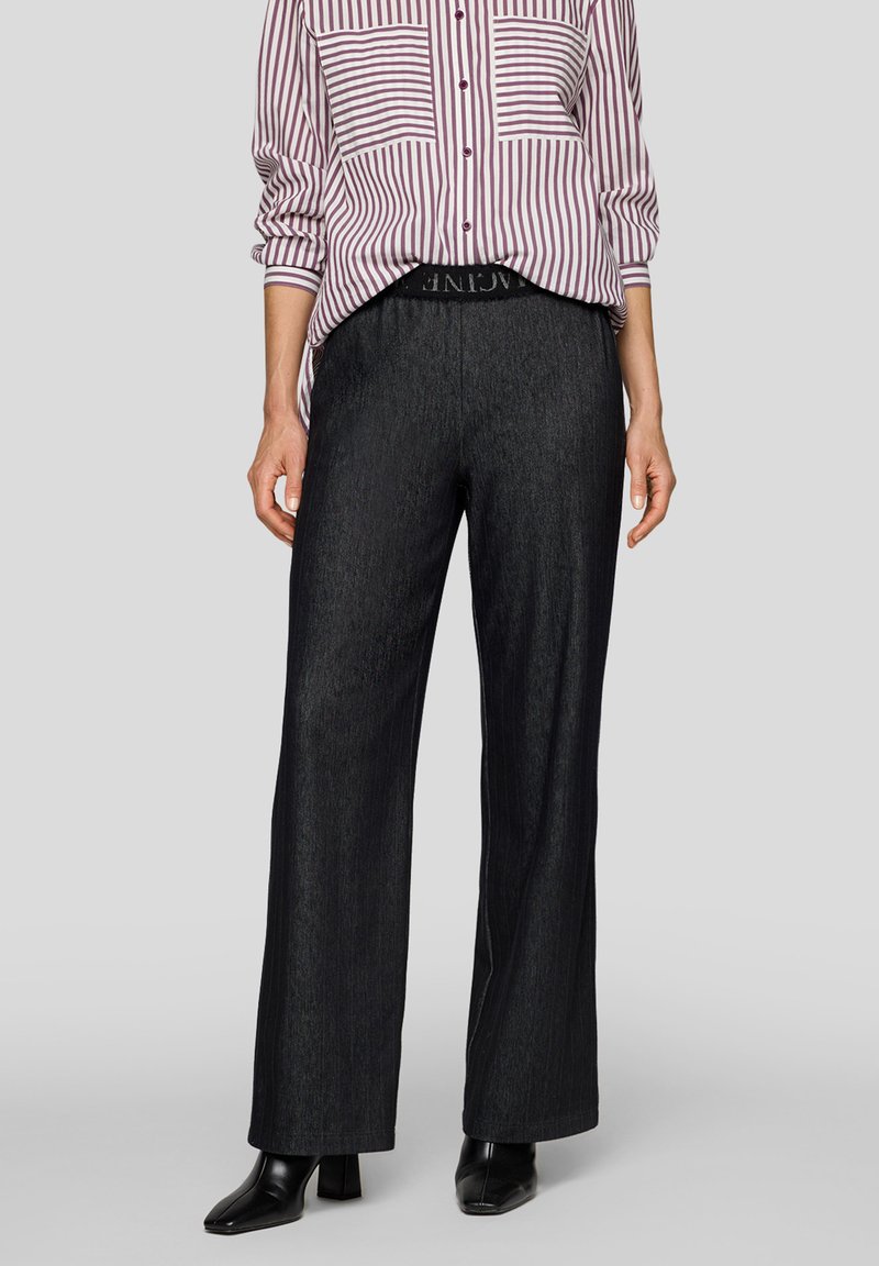 Frau trägt schwarze weite Hosen, schwarze Stiefel mit Absatz und ein lila-weiß gestreiftes Hemd mit hochgekrempelten Ärmeln.