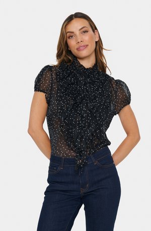 Black sheer blouse with ruffled neckline and short puff sleeves, featuring white polka dots and a flowy texture. Paired with dark jeans.