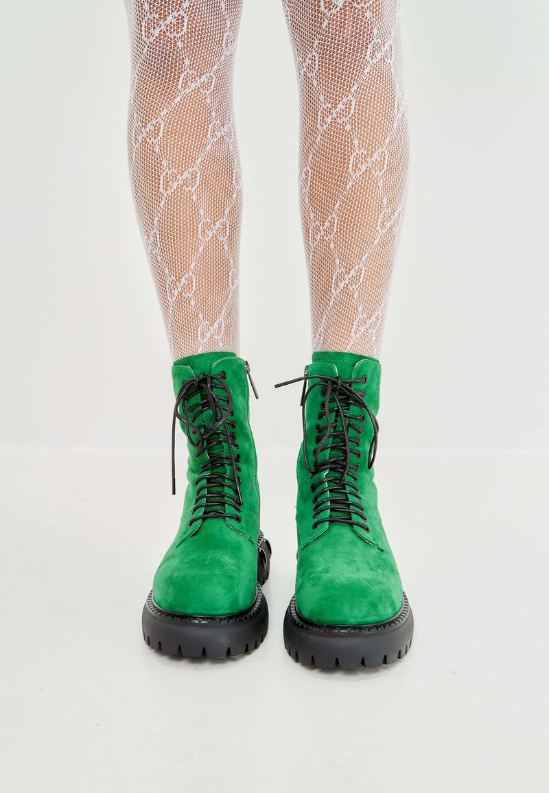 Groene suède enkelboots met zwarte rubberen lugs, met een vetersluiting aan de voorkant, hoge schacht en ronde neus. Benen in witte geprinte visnetkousen.