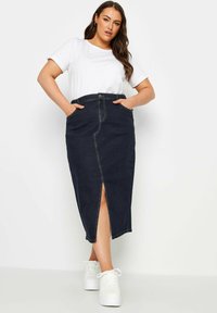 Falda maxi de mezclilla azul oscuro con una abertura frontal, cintura alta, dos bolsillos laterales y detalles de costura amarilla. Combinada con una camiseta blanca y zapatillas.