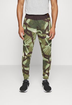 Mand iført joggers med grønne og brune abstrakte camouflage mønstre, sorte talje og sorte træningssko.