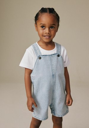 Noor laps punutud juustega, valges T-särgis ja helesinistes teksapükstes, seisab ühevärvilise beeži tausta ees.